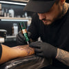 Tattoo artist using a green GTARTISTOO wireless tattoo pen for detailed line work on a client’s arm.