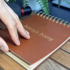 Hand opening a tattoo fake skin book with brown hardcover, gold spiral binding, and synthetic skin practice pages.